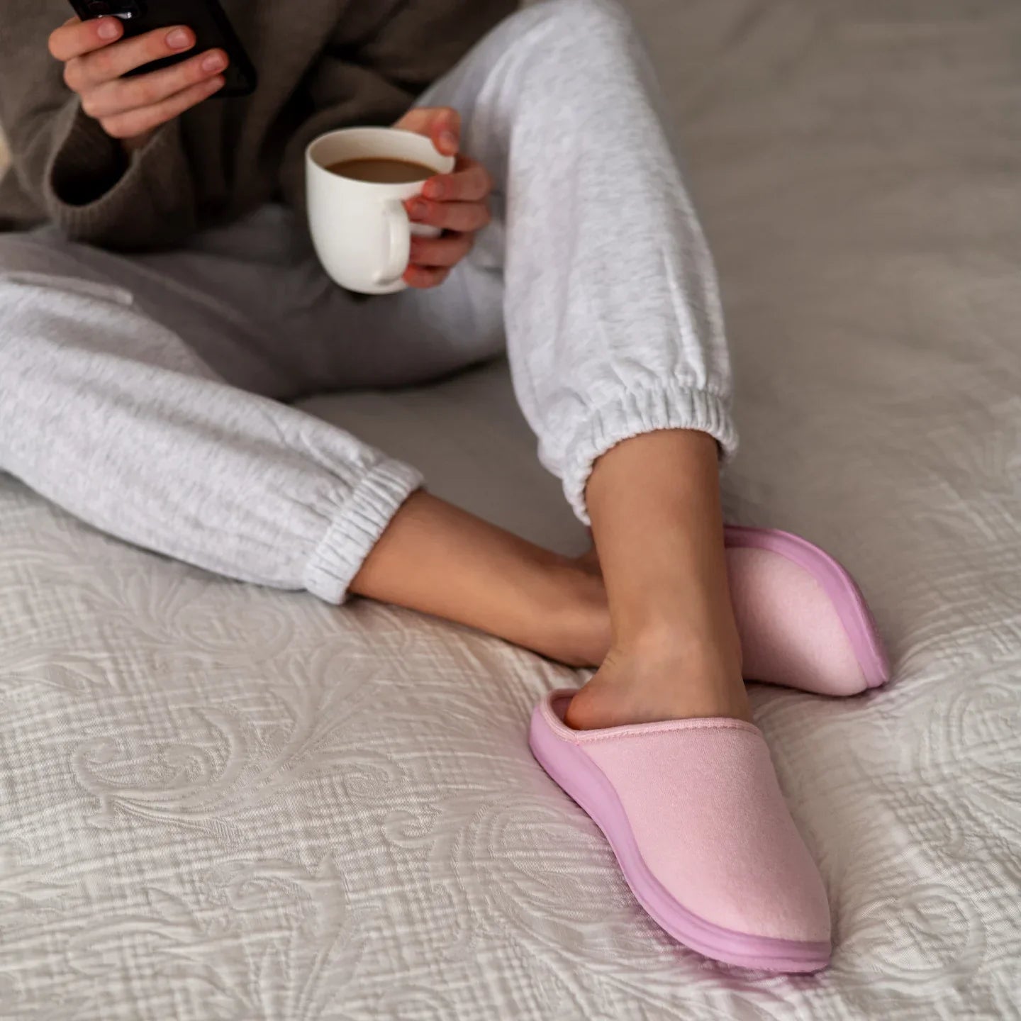 Person relaxing on a bed wearing pink slippers, holding a coffee cup and smartphone.