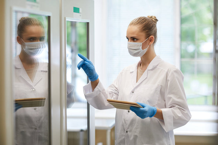 Female healthcare worker in lab coat, mask, and gloves using touchscreen in medical facility