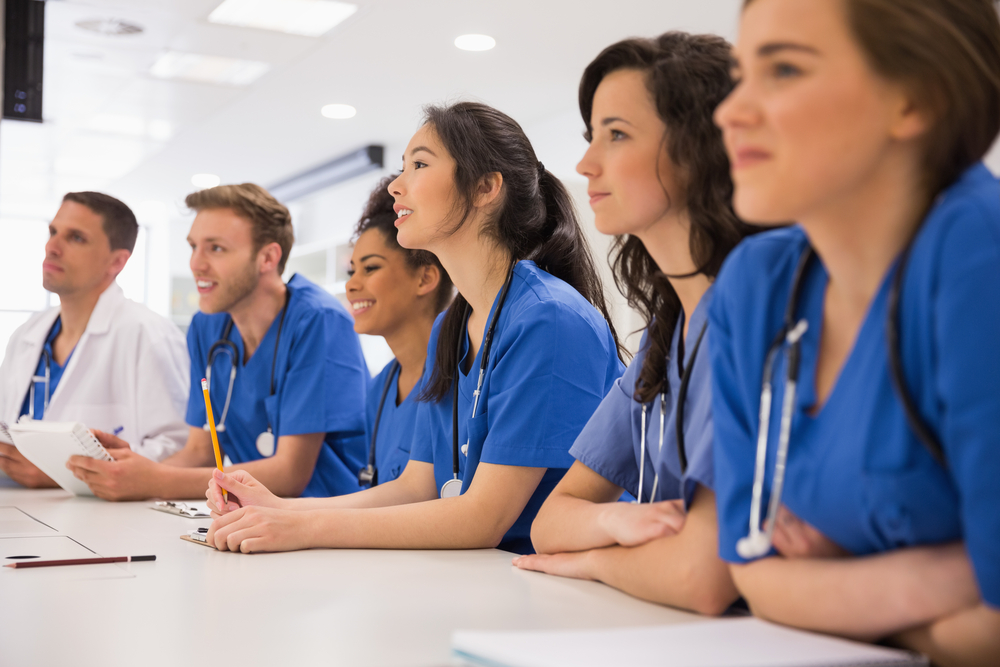 Medical students in blue scrubs attending a classroom lecture with notebooks and stethoscopes.