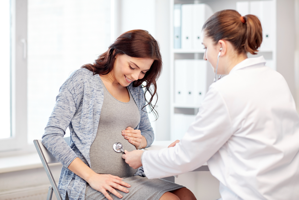 Female doctor using stethoscope on pregnant woman during prenatal checkup in clinic