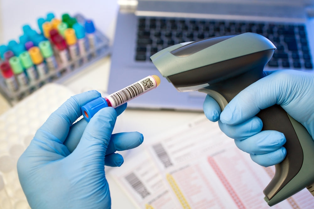Gloved hands scan barcode on blood sample tube in medical lab with test tubes and laptop