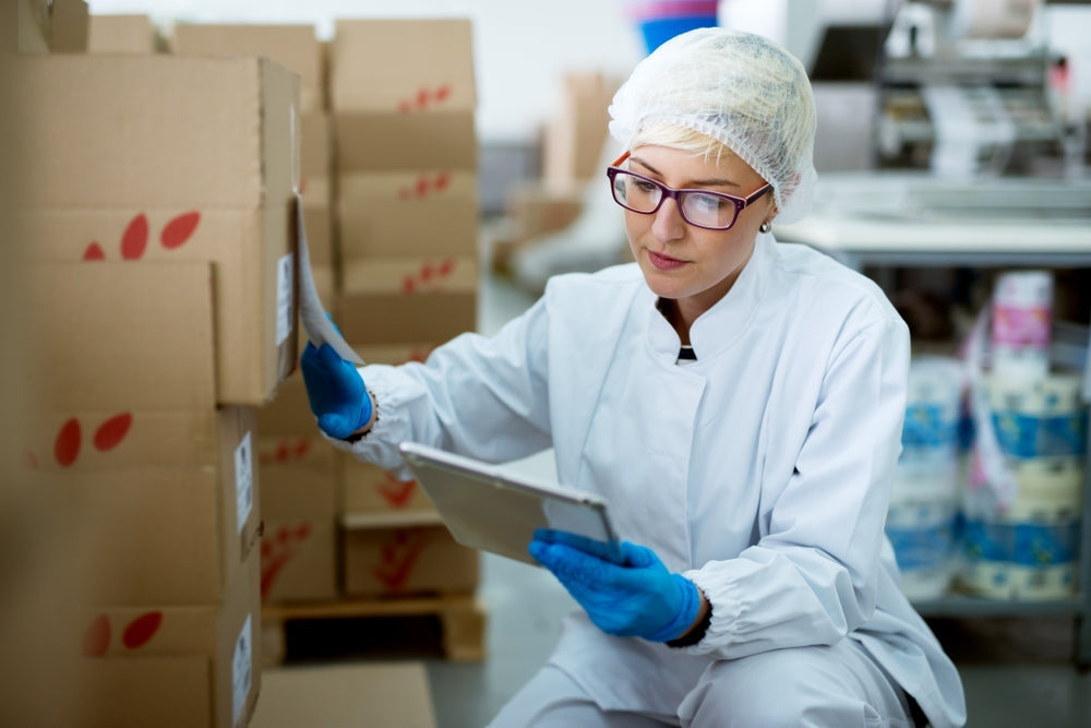 Warehouse worker in medical lab coat and gloves checking inventory on tablet near stacked boxes
