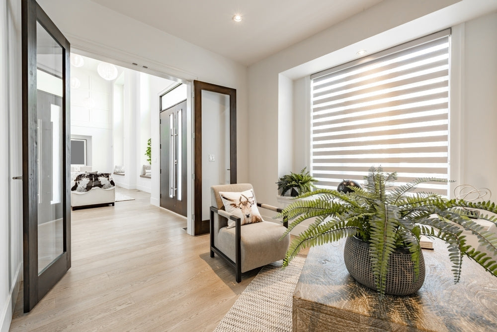 Modern home office with armchair, large window blinds, indoor plants, and light wood flooring