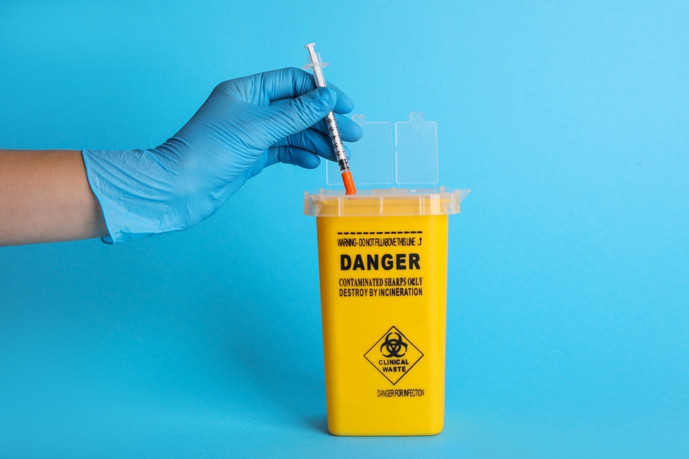 Gloved hand disposing syringe into yellow sharps bin for clinical waste on blue background