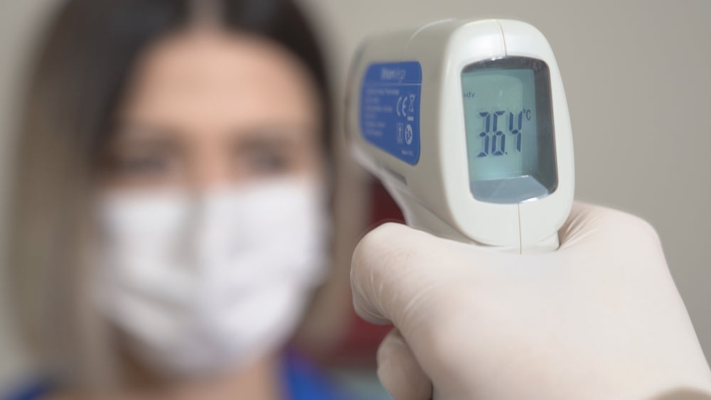 Gloved hand using digital infrared forehead thermometer to check temperature of masked woman